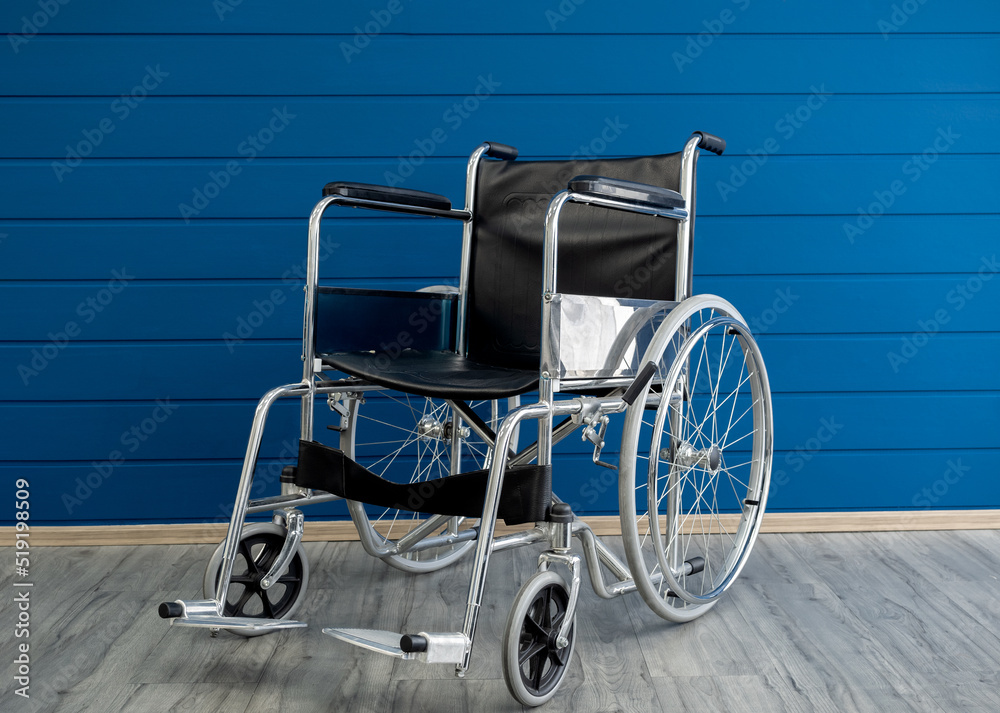 Empty steel wheelchair with black leather seat and backrest on the blue wooden plank wall background. A chair with wheels for use as a transport by a person who unable to walk, illness, injury.
