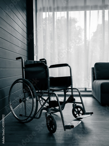 Empty black wheelchair near the blue wall and grey with white curtain at glass window in the living room vertical style, waiting for patient services. Lonely wheelchair with nobody in hospital room.