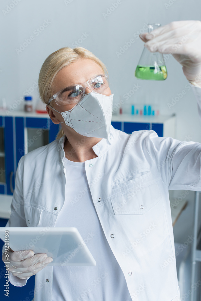 Scientist in goggles looking at blurred flask and holding digital tablet in lab. Scientist in goggles looking at blurred flask and holding digital tablet in lab.
