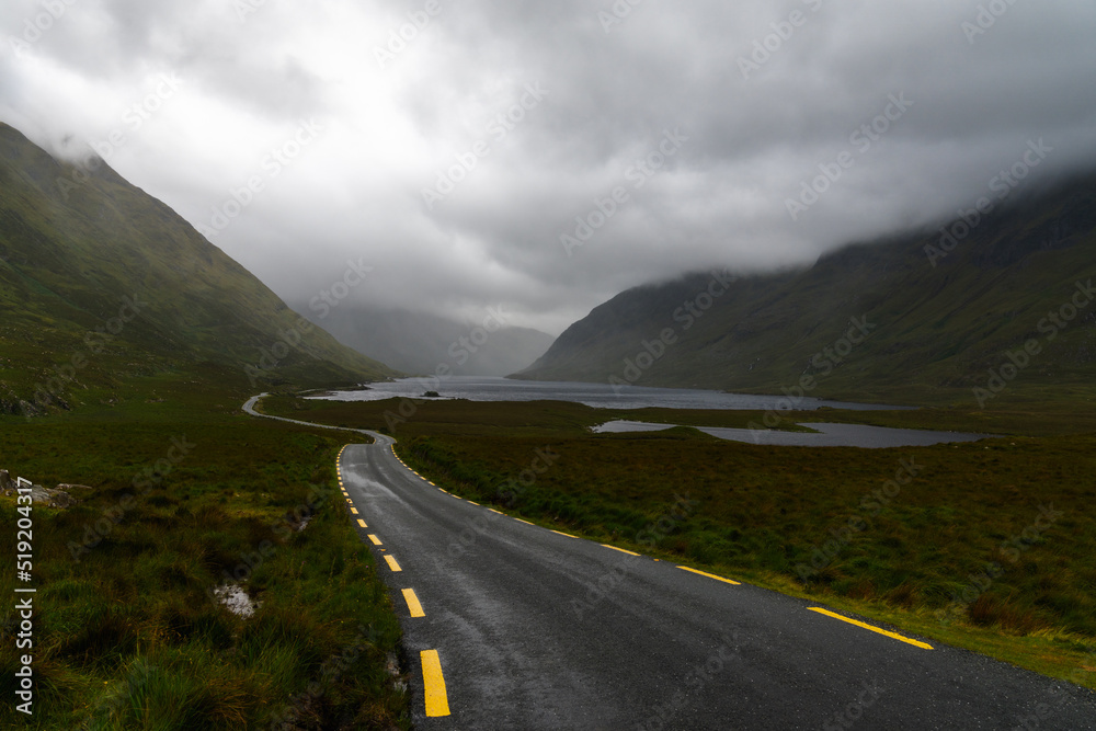 Naklejka premium narrow black-top highway leads through an overcast mountain valley with fog and mist and lakes o nthe valley bottom