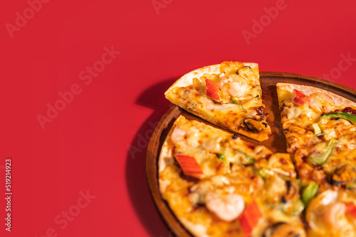 Close up slice of tasty hot baked seafood crispy pizza - mussels, shrimps and kani with chili pepper and melted mozzarella cheese on round pizza dough, on a wooden pizza pan over the red background.