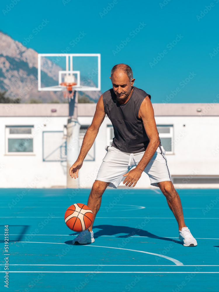 Fototapeta premium Portrait of Turkish Cypriot elderly athletic man having fun playing basketball with a cute dog jack russel terrier on blue color playground outdoor at summer sunny day