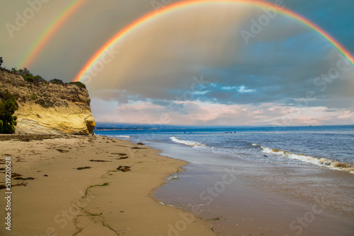 a gorgeous summer landscape at the beach with rocky cliffs covered in lush green trees and plants, blue ocean water, surfers in the water and blue sky, clouds and a rainbow at Leadbetter Beach