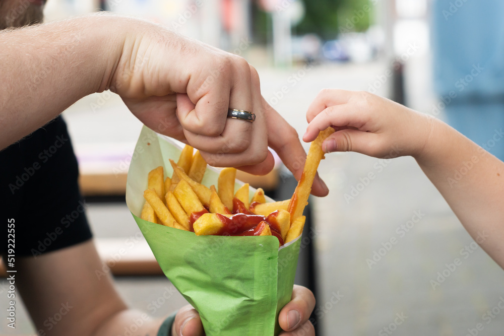 Big hand of a grown-up man and the little hand of a kid reaching for some Belgian fries with ketchup from a street food truck. Family fast food outside.