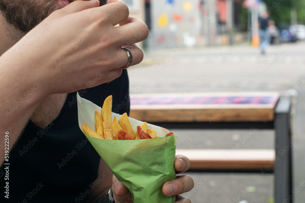 Man eating some street food outside - french fries with ketchup in a paper cone. Caucasian bearded guy having fast food.