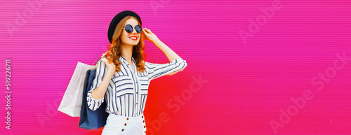 Portrait of beautiful smiling young woman with shopping bags wearing white striped shirt, summer black round straw hat on pink background, blank copy space for advertising text