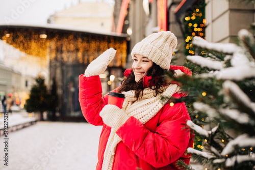 Young pretty brunette woman in red jacket, white hat, scarf and mittens with cap of coffee to go