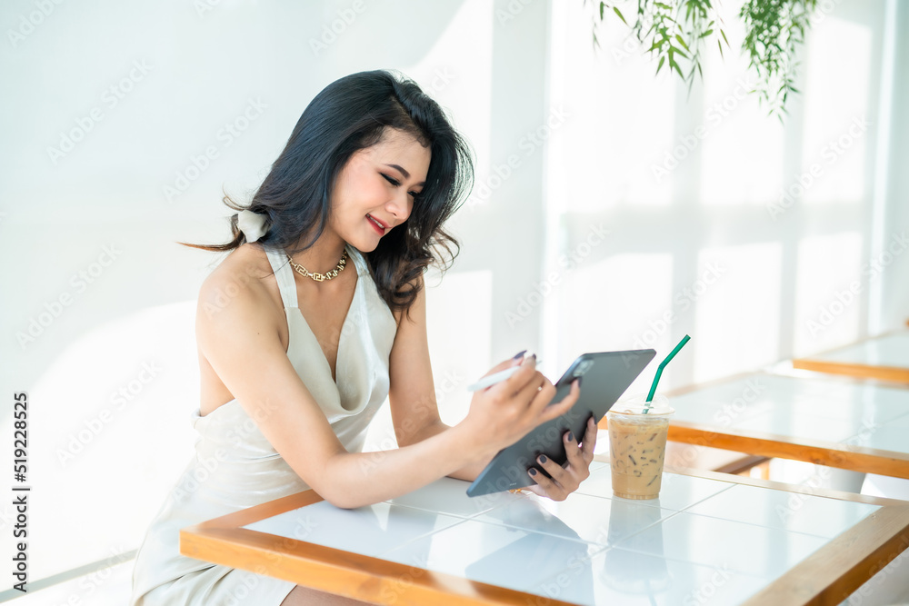 Obraz premium beautiful asian freelance Success people business woman fashion model in summer white sleeveless dress using casual working with tablet for browsing internet, chatting and blogging in coffee shop.