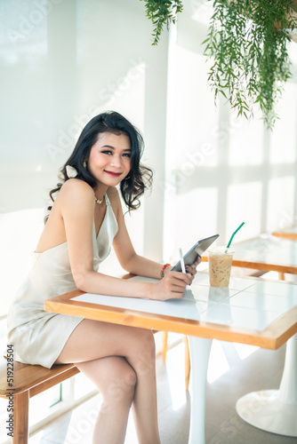 beautiful asian freelance Success people business woman fashion model in summer white sleeveless dress using casual working with tablet for browsing internet, chatting and blogging in coffee shop. © Thinapob