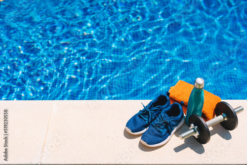 sport accessories in front of a swimming pool. orange towel, blue water bottle, weights. background of a swimming pool in a garden. © A&NStudio
