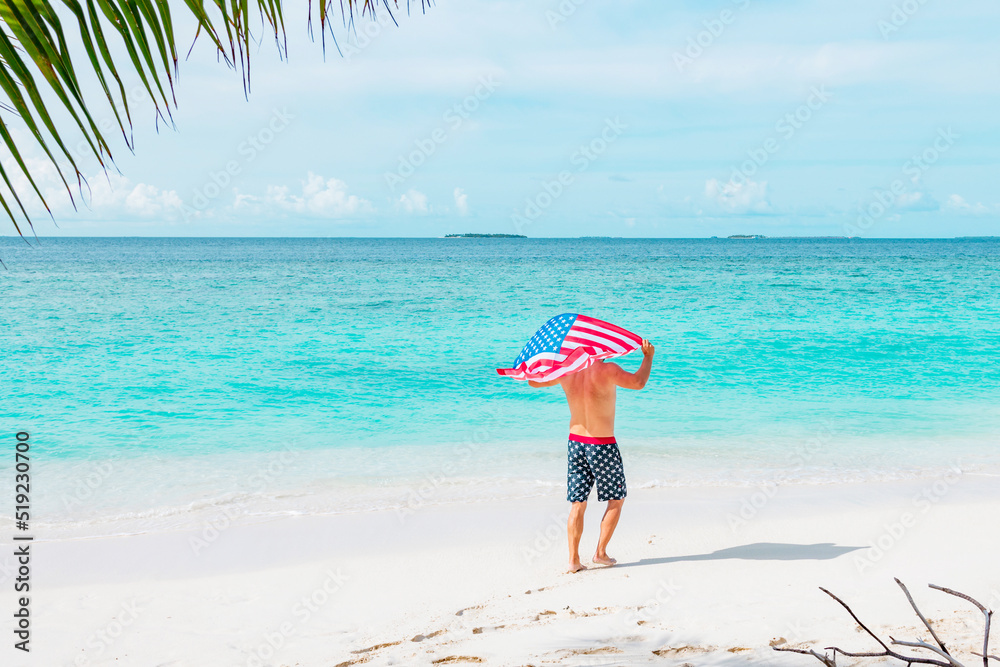 young athletic man stands in swimming trunks with an American flag on his shoulders near ocean sea shore, concept of celebrating America's USA Independence Day on July 4 and patriotic education, sense young athletic man stands in swimming trunks with an American flag on his shoulders near ocean sea shore, concept of celebrating America's USA Independence Day on July 4 and patriotic education, sense