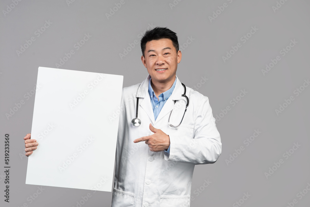 Smiling handsome adult asian male doctor in white coat with stethoscope points finger at banner Smiling handsome adult asian male doctor in white coat with stethoscope points finger at banner