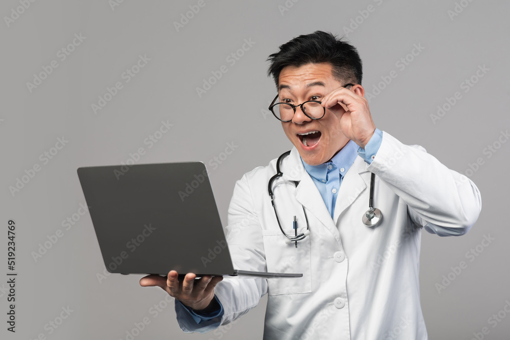Shocked cheerful millennial asian man doctor in white coat with open mouth takes off glasses, looks at laptop Shocked cheerful millennial asian man doctor in white coat with open mouth takes off glasses, looks at laptop