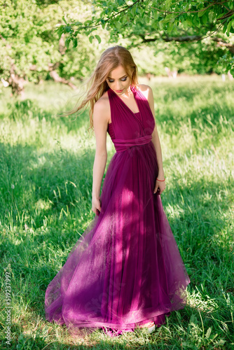 Bridesmaid stands in the summer garden and demonstrates a festive dress of purple color, posing looking down. Professional makeup and hair styling, High quality photo