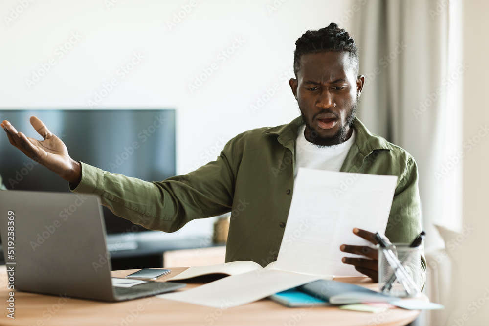 Shocked black man reading paper letter mail and spreading hands, sitting at table with laptop at home office Shocked black man reading paper letter mail and spreading hands, sitting at table with laptop at home office