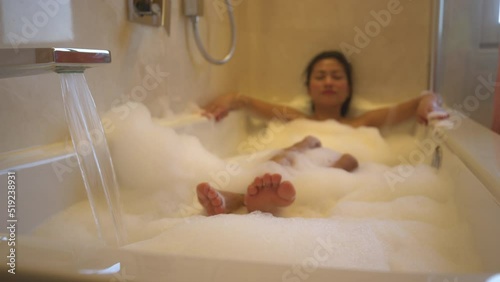 Water flowing from tap into bath and young lady relaxing in bathtub full of foam. Relaxation and spa treatment in home bathroom for the end of the day. Young lady treating herself with bubble bath.