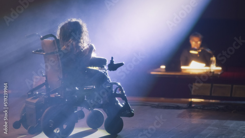 A woman with spinal muscular atrophy having an audition on a theater stage illuminated by a spotlight with a director of the performance