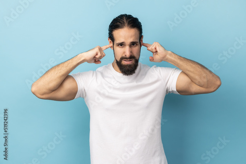 I don't want to here this. Portrait of man with beard wearing white T-shirt standing and holding fingers on his ear, unpleasant sounds. Indoor studio shot isolated on blue background.