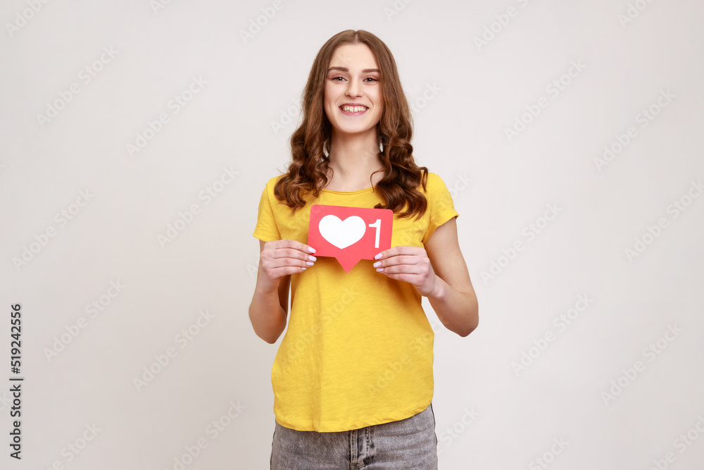 Like button of social media. Portrait of pleasant looking young woman with wavy hair holding heart icon, recommending to follow and share blog. Indoor studio shot isolated on gray background. Like button of social media. Portrait of pleasant looking young woman with wavy hair holding heart icon, recommending to follow and share blog. Indoor studio shot isolated on gray background.