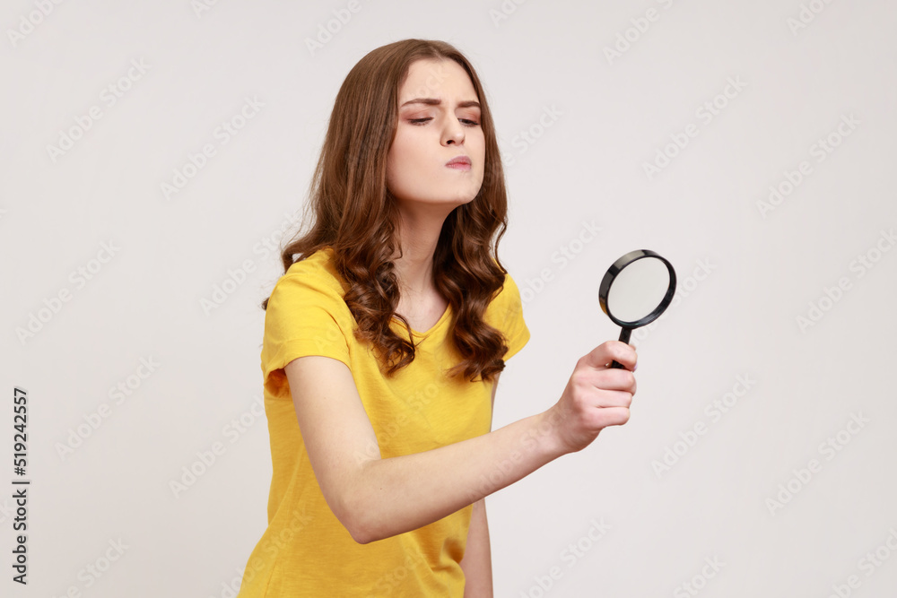 Young attractive curious female with brown hair looking through magnifying glass, spying, finding out something, inspecting crime scene. Indoor studio shot isolated on gray background. Young attractive curious female with brown hair looking through magnifying glass, spying, finding out something, inspecting crime scene. Indoor studio shot isolated on gray background.