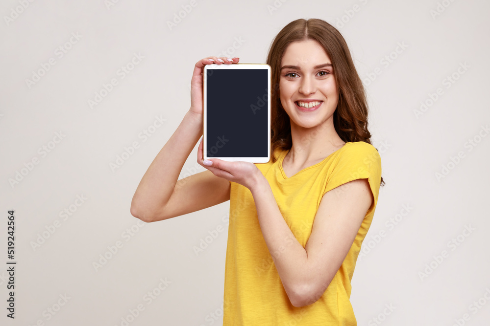 Portrait of beautiful smiling young teenage with brown hair standing and holding tablet empty screen, sowing device and looking at camera. Indoor studio shot isolated on gray background. Portrait of beautiful smiling young teenage with brown hair standing and holding tablet empty screen, sowing device and looking at camera. Indoor studio shot isolated on gray background.