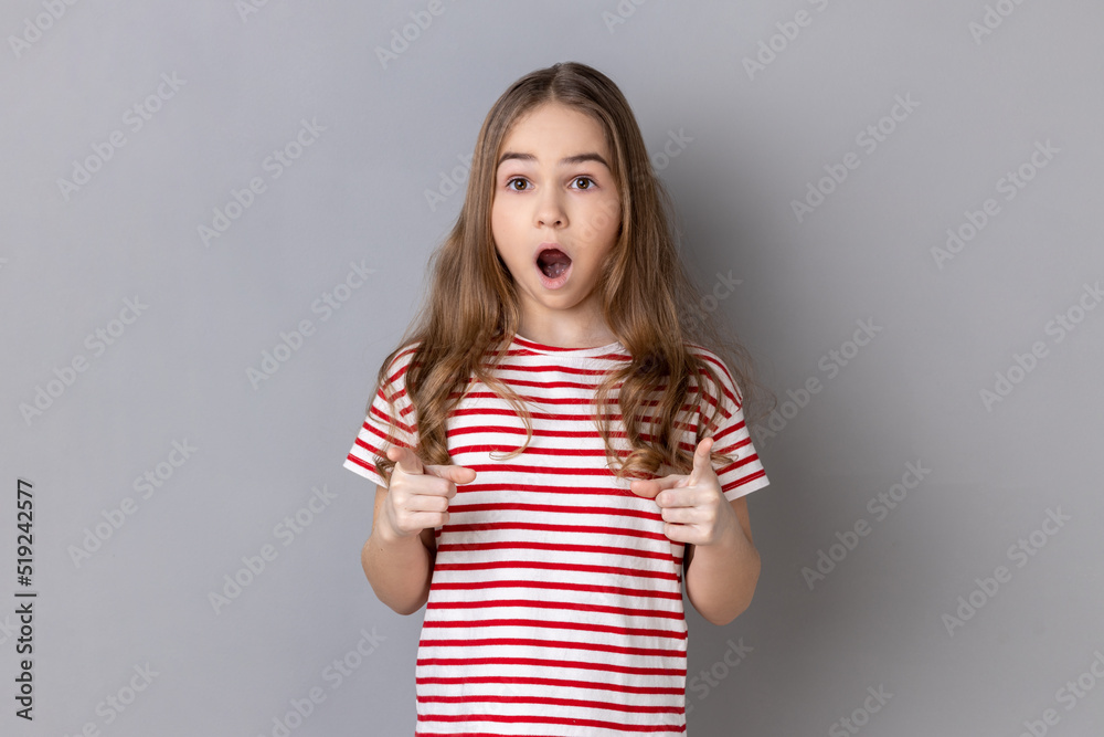 Portrait of astonished little girl wearing striped T-shirt pointing to camera with surprised shocked expression and open mouth, making choice. Indoor studio shot isolated on gray background.