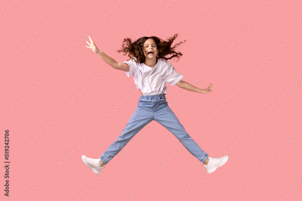 Full length portrait of excited adorable little girl wearing white T-shirt jumping in air and spread arms, smiling happily, having fun. Indoor studio shot isolated on pink background.
