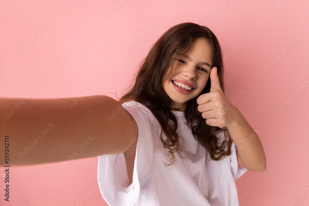 Little girl wearing white T-shirt has optimistic face expression, making selfie, expresses sincere emotions, showing thumb up, point of view photo. Indoor studio shot isolated on pink background. Little girl wearing white T-shirt has optimistic face expression, making selfie, expresses sincere emotions, showing thumb up, point of view photo. Indoor studio shot isolated on pink background.