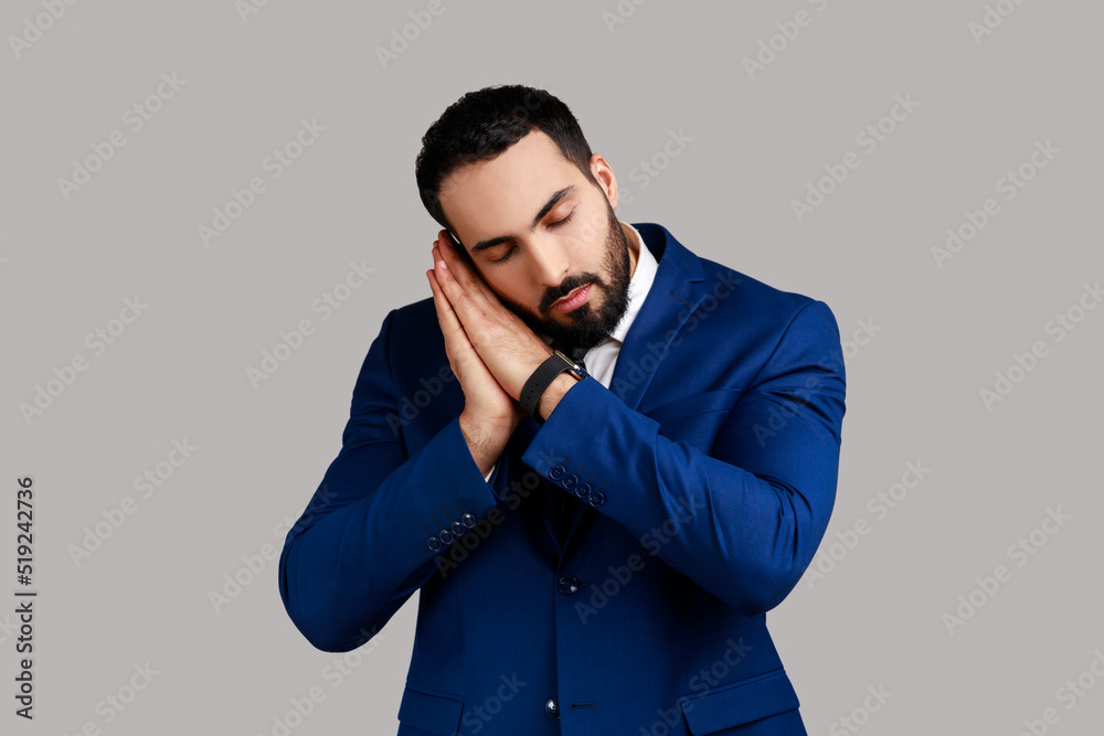 Portrait of tired bearded man sleeping laying down on her palms, keeping eyes closed, wearing white sweater, wearing official style suit. Indoor studio shot isolated on gray background. Portrait of tired bearded man sleeping laying down on her palms, keeping eyes closed, wearing white sweater, wearing official style suit. Indoor studio shot isolated on gray background.