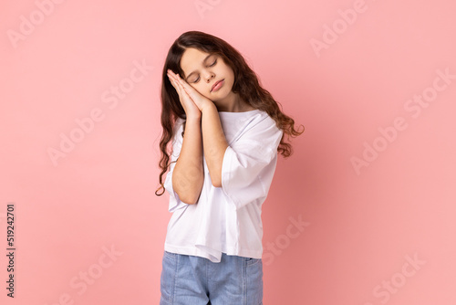 Portrait of cute little girl wearing white T-shirt sleeping laying down on her palms, having comfortable nap and resting, dozing off. Indoor studio shot isolated on pink background.