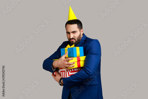 Fotografiet Sad bearded man with party cone on his head holding stack of presents in hands, being greedy to share gifts, wearing official style suit