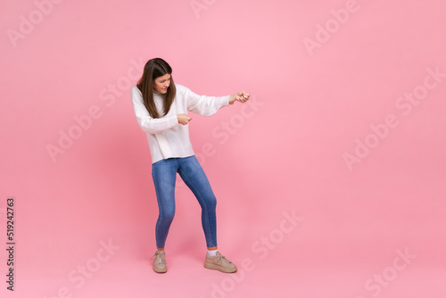 Full length portrait of girl pulling invisible rope with effort, using all strength to achieve goal, wearing white casual style sweater. Indoor studio shot isolated on pink background.