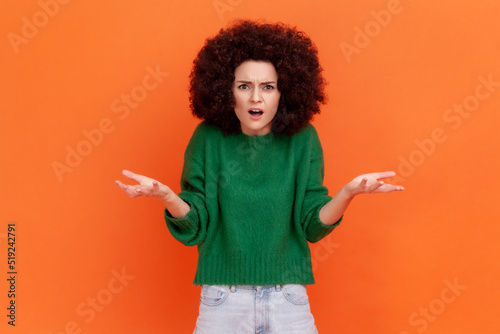 What do you want? Woman with Afro hairstyle wearing green casual sweater asking who why make this conflict, looking with annoyed indignant expression. Indoor studio shot isolated on orange background.
