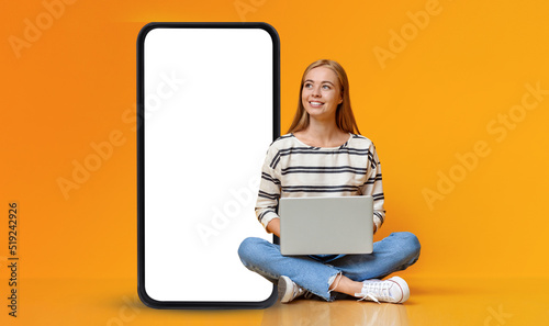 Teenage girl sitting with laptop on floor, looking at smartphone