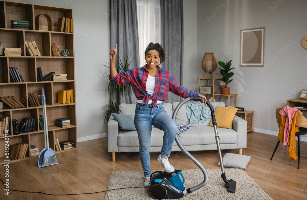 Cheerful millennial black housewife vacuuming with vacuum cleaner and pointing finger up at empty space