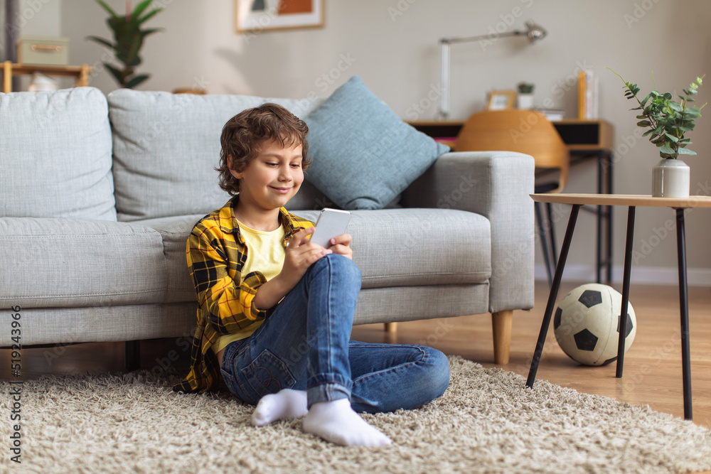 Cute little boy web surfing online on smartphone, watching video or playing games, sitting on floor at home, empty space Cute little boy web surfing online on smartphone, watching video or playing games, sitting on floor at home, empty space