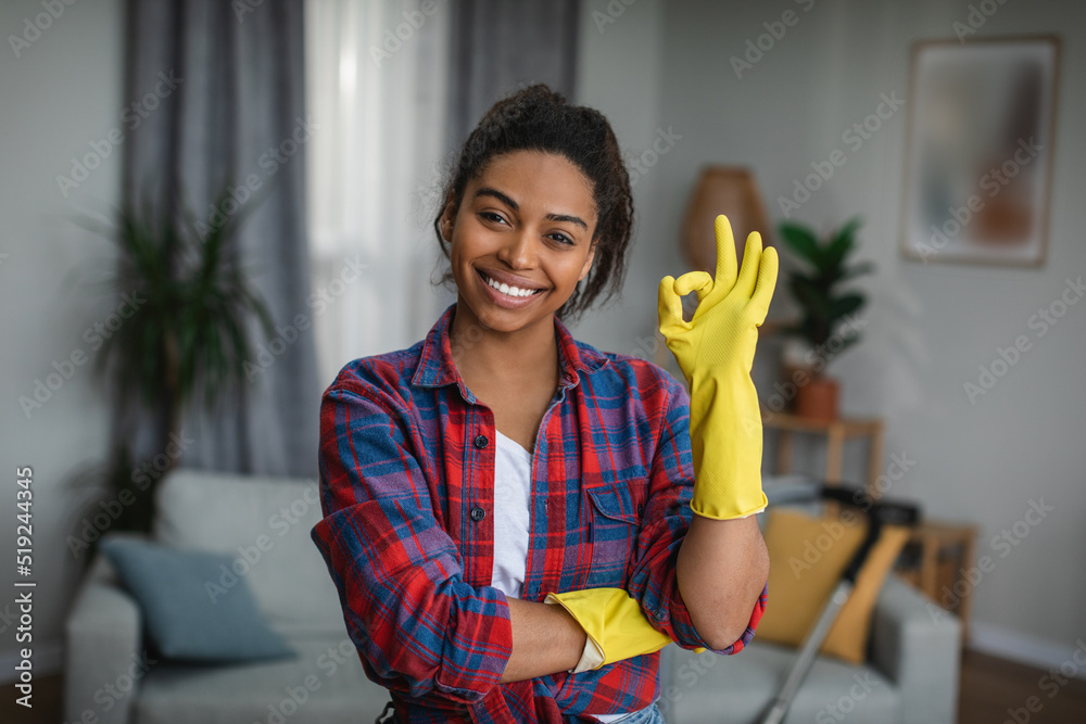 Glad millennial black lady in rubber gloves shows ok sign with hand in living room interior Glad millennial black lady in rubber gloves shows ok sign with hand in living room interior