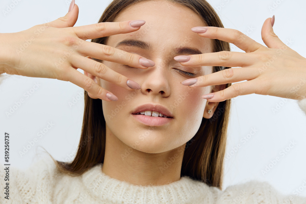 Fototapeta premium a beautiful, sophisticated woman in a white sweater stands on a gray background and covers her face with her fingers with a perfect nude manicure. Horizontal Studio Photography