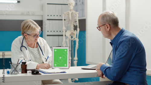 General practitioner vertically holding tablet with greenscreen on display in medical cabinet. Mockup template with blank chroma key background and isolated copyspace. Tripod shot.