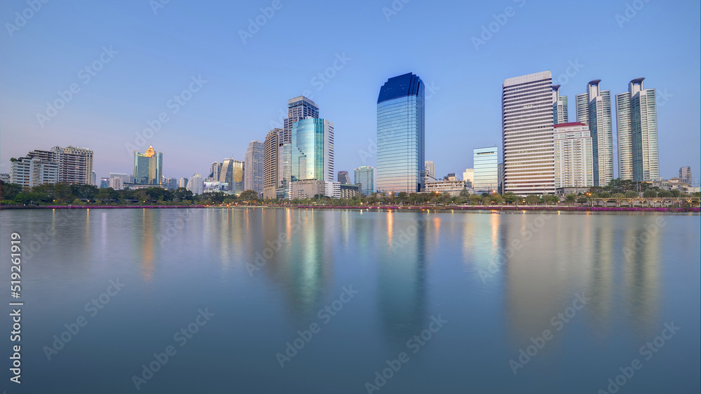 Night skyline of modern lakeside skyscrapers with glass curtain walls and dazzling city lights reflected in the smooth lake water in beautiful Benjakiti Park at blue dusk, in Bangkok, Thailand, Asia