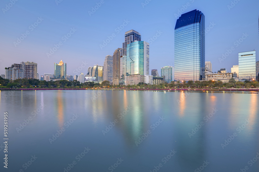 Night skyline of modern lakeside skyscrapers with glass curtain walls and dazzling city lights reflected in the smooth lake water in beautiful Benjakiti Park at blue dusk, in Bangkok, Thailand, Asia