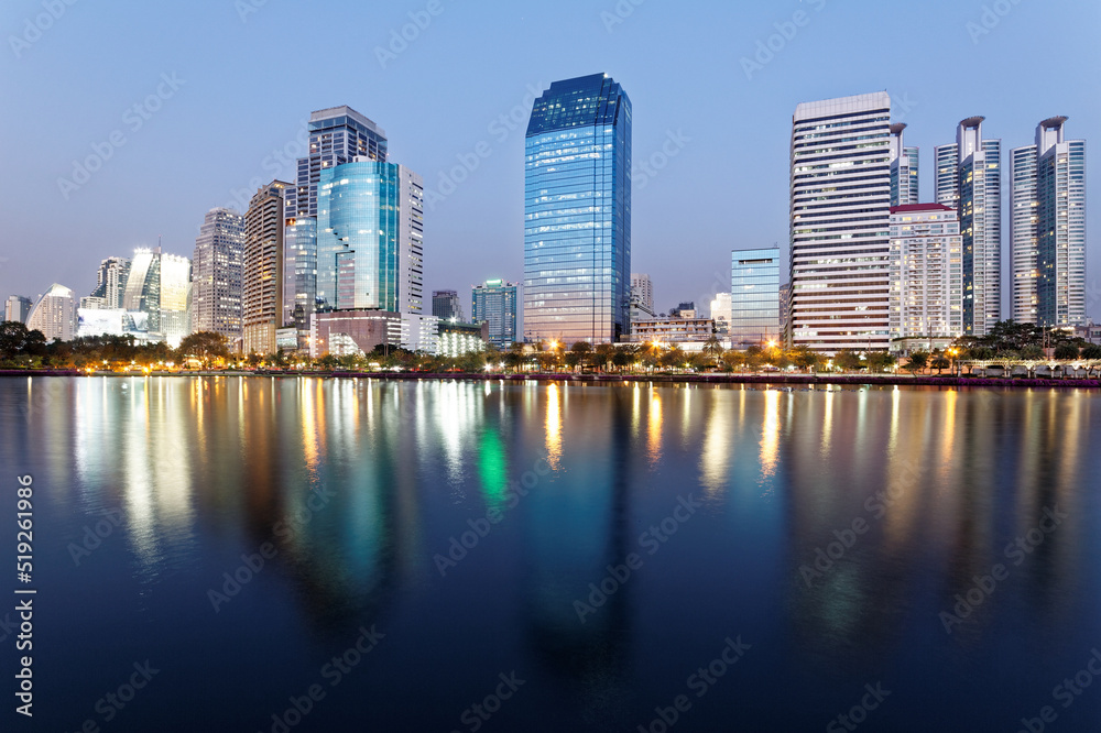 Night skyline of modern lakeside skyscrapers with glass curtain walls and dazzling city lights reflected in the smooth lake water in beautiful Benjakiti Park at blue dusk, in Bangkok, Thailand, Asia