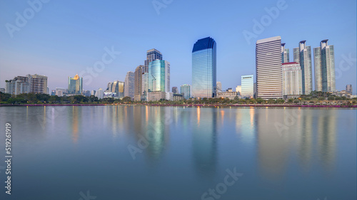 Night skyline of modern lakeside skyscrapers with glass curtain walls and dazzling city lights reflected in the smooth lake water in beautiful Benjakiti Park at blue dusk  in Bangkok  Thailand  Asia
