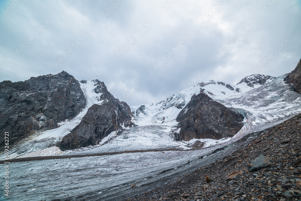 Dramatic landscape with two glacier icefalls on large snow mountain range with sharp rocks under gray cloudy sky. Long glacier with icefall in high altitude. Gloomy scenery in mountains in overcast.