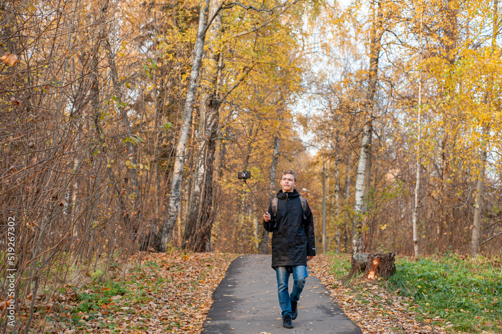 A teenage boy walks through an autumn park and shoots a video. A Caucasian boy in a black jacket holds a monopod with a smartphone. Blogger. Emotional communications..