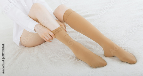 Beautiful woman putting on stocking indoors, closeup. Beautiful long female legs in stockings. Girl putting on stockings at home in a white room. Beige knee socks or socks photo