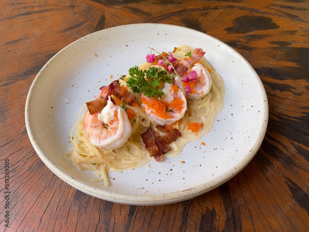 Spaghetti carbonara cream sauce with prawn, shrimp roe and crispy bacon decorated with edible flowers and parsley, served in white ceramic dish on wooden table. Selective focus
