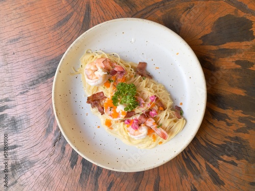 Spaghetti carbonara cream sauce with prawn, shrimp roe and crispy bacon decorated with edible flowers and parsley, served in white ceramic dish on wooden table. Selective focus photo