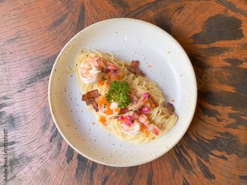 Spaghetti carbonara cream sauce with prawn, shrimp roe and crispy bacon decorated with edible flowers and parsley, served in white ceramic dish on wooden table. Top view photo