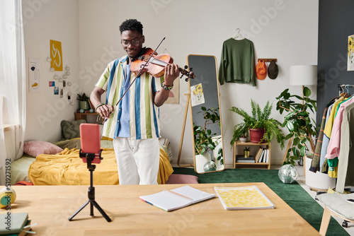 African young blogger standing in front of smartphone on tripod and playing violin for his followers during online streaming photo